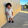 Azariah joined the competition — help win amazing prizes! toddler, child, curly_hair, floral_clothing, shorts, shirt, standing, wall, outdoor, sidewalk, daylight, shadow, curious_expression, footwear, casual_pose, sunlight, texture, blue_sky, residential_area, young_child