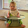 baby, child, smiling, high_chair, table, bib, green_clothing, pony_tail, indoor, window_blinds, wooden_table, happy, seated, infant, cute, person, modern_furniture, natural_light, home, portrait