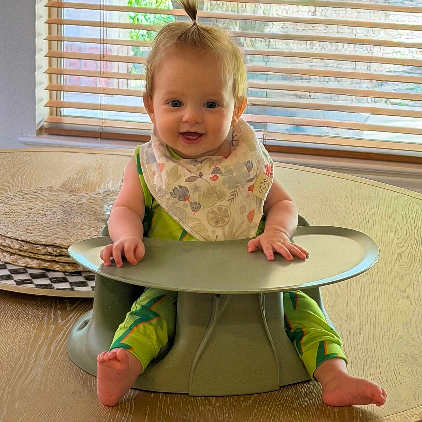 Lucy is registered to the contest to win money with this photo: baby, child, smiling, high_chair, table, bib, green_clothing, pony_tail, indoor, window_blinds, wooden_table, happy, seated, infant, cute, person, modern_furniture, natural_light, home, portrait