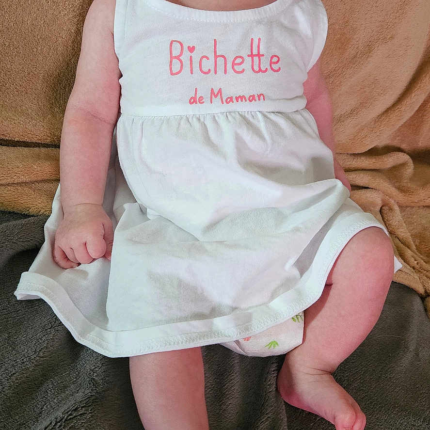 Alexie a rejoint le concours — aidez-le/la à gagner de superbes lots ! baby, blanket, child, clothing, cozy, curious, cute, face, feet, hand, indoor, infant, pink_text, portrait, sitting, skin, soft_texture, white_dress, white_hat, young_child