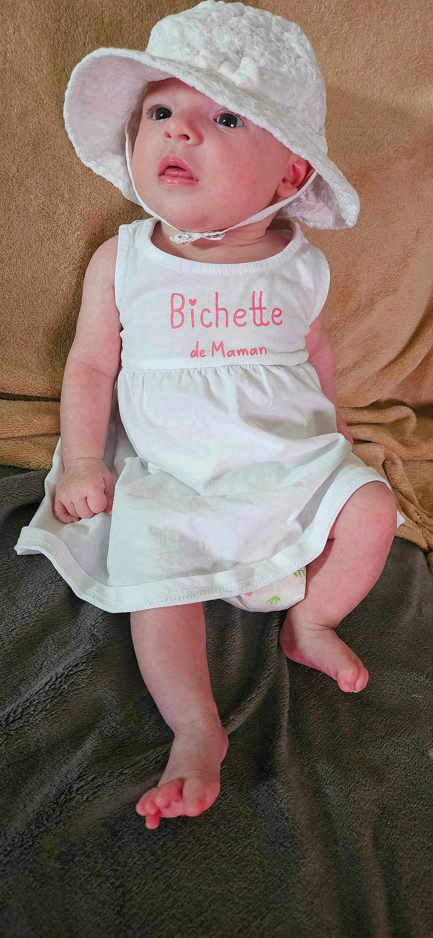 Alexie a rejoint le concours — aidez-le/la à gagner de superbes lots ! baby, child, infant, white_hat, white_dress, pink_text, blanket, sitting, curious, face, feet, hand, indoor, soft_texture, cute, portrait, young_child, cozy, skin, clothing