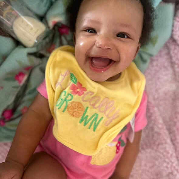Janiyla joined the competition — help win amazing prizes! baby, smiling, happy, child, infant, bib, pink_clothing, yellow_bib, blanket, soft, indoors, cute, joyful, portrait, sitting, face, human, young_child, expressive, cheerful