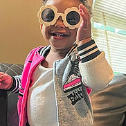 Janiyla is registered to the contest to win money with this photo: blinds, child, clothing, couch, curly_hair, cute, fashion, hair, happy, indoor, jacket, light, person, playful, portrait, smile, sunglasses, toddler, white_shirt, window