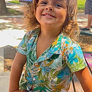 Sebastian is registered to the contest to win money with this photo: child, smiling, curly_hair, outdoor, park, tropical_shirt, floral_pattern, toy, purple_toy, sunlight, bench, person, sidewalk, tree, greenery, casual_clothing, happy, playful, daytime, portrait
