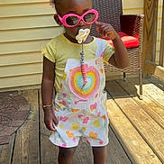 Jaior Milani is registered to the contest to win money with this photo: toddler, child, sunglasses, pacifier, overalls, heart_pattern, yellow_shirt, pink_shoes, bows, bracelet, wooden_deck, porch, chair, outdoor, sunlight, fashion, cute, standing, person, summer