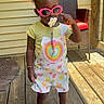 toddler, child, sunglasses, pacifier, overalls, heart_pattern, yellow_shirt, pink_shoes, bows, bracelet, wooden_deck, porch, chair, outdoor, sunlight, fashion, cute, standing, person, summer