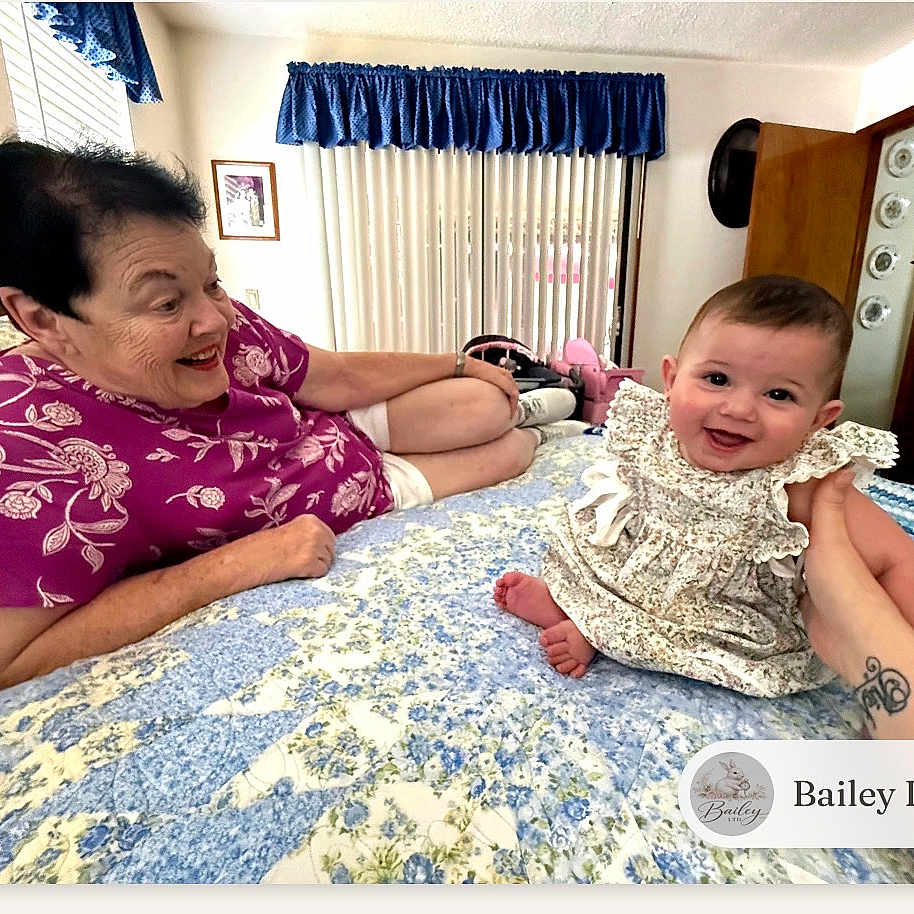 Bailey joined the competition — help win amazing prizes! baby, elderly_woman, bed, quilt, smiling, indoor, hand, floral_dress, purple_shirt, window, curtains, furniture, drawer, person, happy, home, tattoo, portrait, family, support