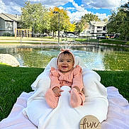 Abby joined the competition — help win amazing prizes! baby, infant, smiling, pink_clothing, knitted_outfit, headband, cushion, blanket, grass, outdoor, pond, trees, blue_sky, clouds, wooden_sign, five_months, happy, child, portrait, nature