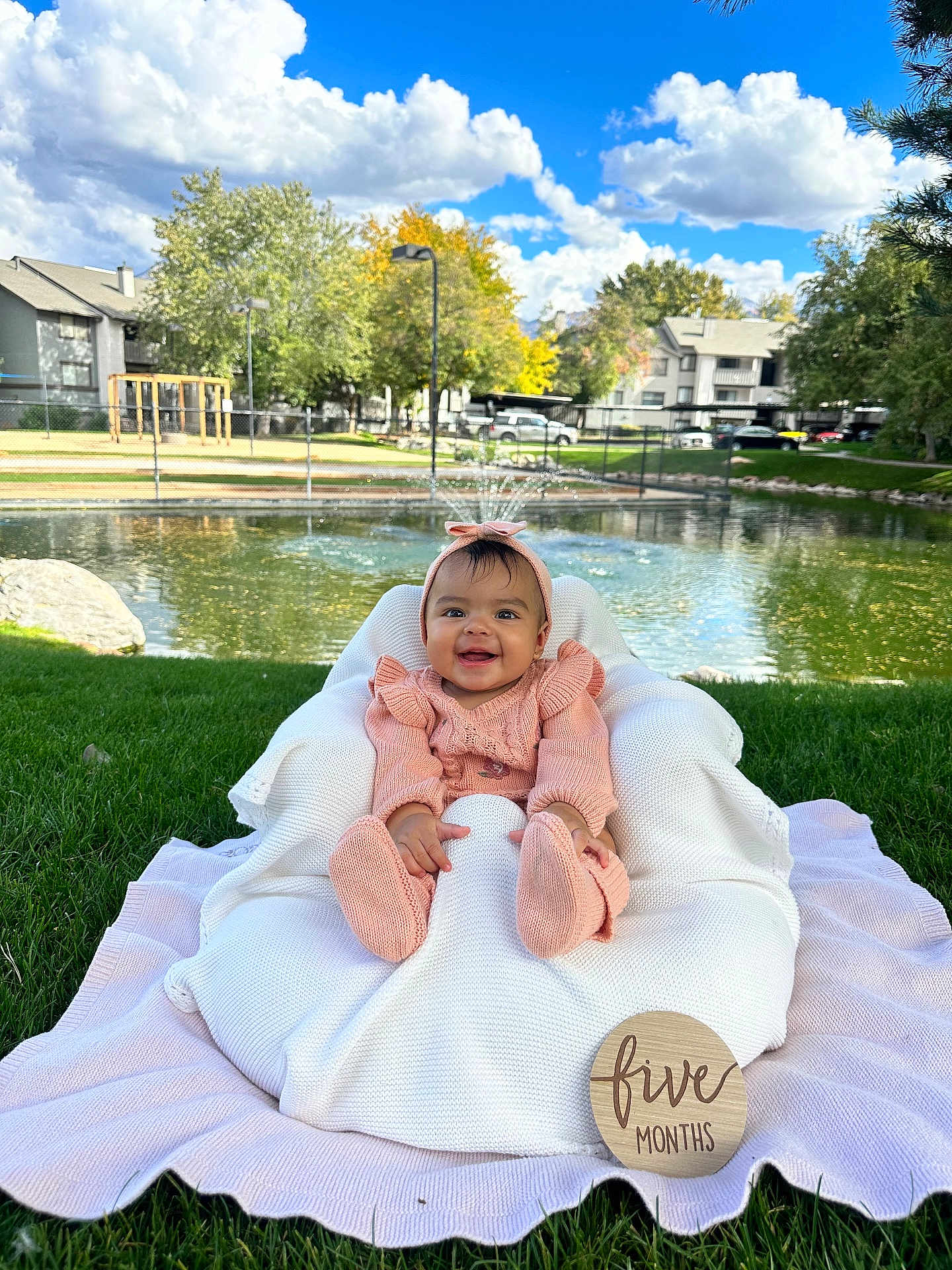 Abby joined the competition — help win amazing prizes! baby, infant, smiling, pink_clothing, knitted_outfit, headband, cushion, blanket, grass, outdoor, pond, trees, blue_sky, clouds, wooden_sign, five_months, happy, child, portrait, nature