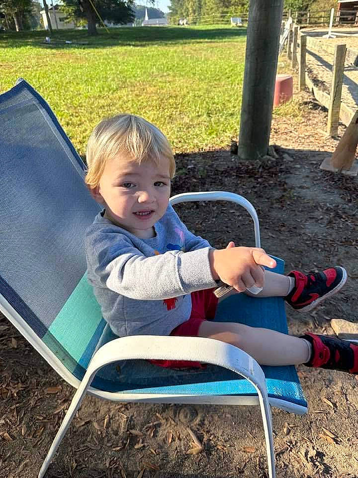 Koda is registered to the contest to win money with this photo: chair, child, comfort, fun, grass, green, human_leg, lap, leisure, outdoor_furniture, people_in_nature, person, plant, recreation, shade, shorts, sitting, sock, thigh, toddler