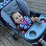baby, stroller, child, toy, wooden_deck, leaves, pink_clothing, floral_pattern, sneakers, seat_belt, outdoor, happy, smiling, infant, person, face, hand, footwear, playful, daylight