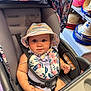 accessory, baby, bib, child, clothing, colorful, cute, expression, face, floral_pattern, hat, indoors, infant, person, portrait, seatbelt, shopping, sitting, store, stroller