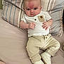 baby, infant, bed, pillow, clothing, white_shirt, beige_pants, white_shoes, face, person, cute, indoors, young_child, patterned_pillow, resting, lying_down, casual, comfortable, expression, small