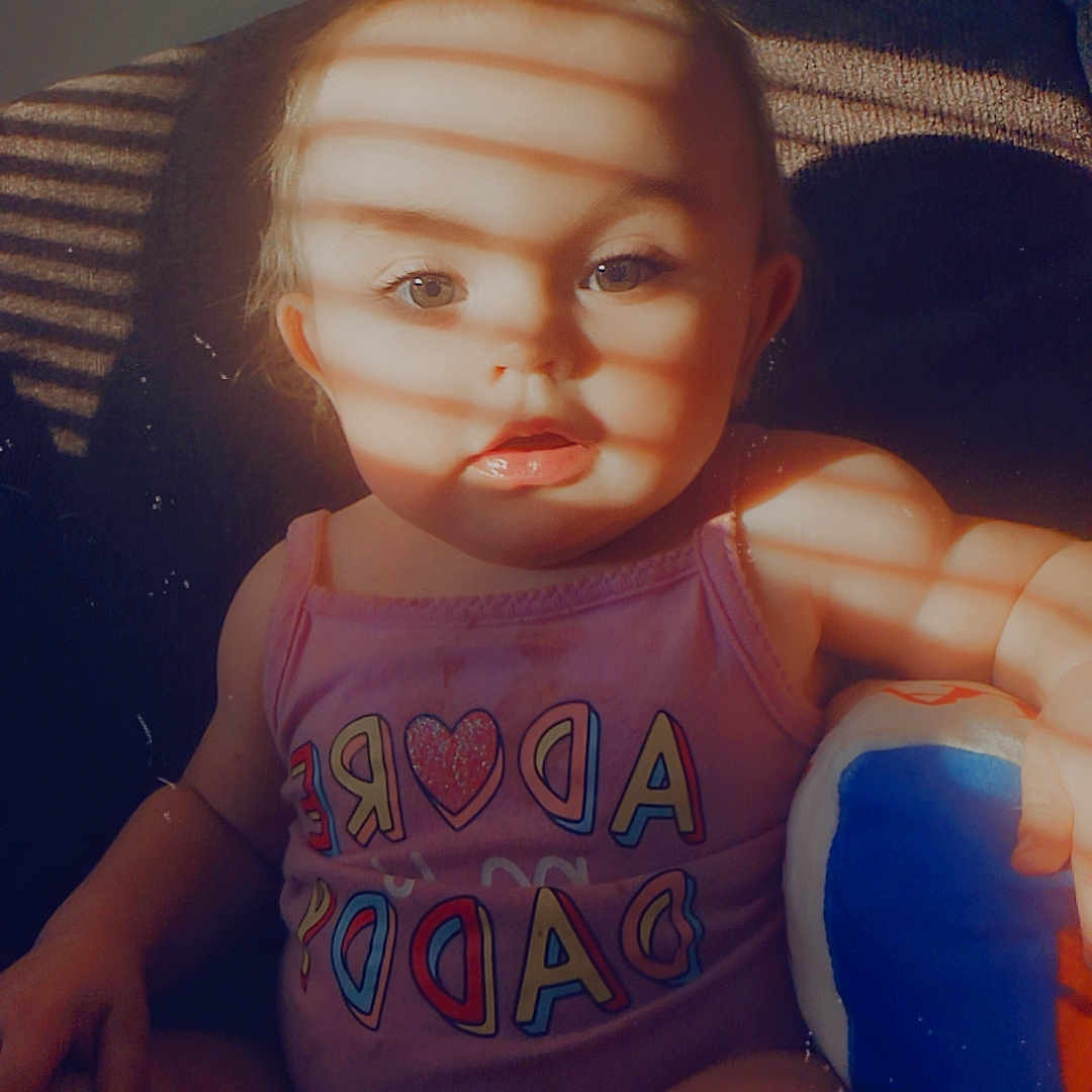Maybrie is registered to the contest to win money with this photo: baby, child, infant, sitting, portrait, face, eyes, expression, pink_clothing, romper, toy, blue_toy, couch, indoor, window_light, sunlight, shadows, striped_shadows, close_up, photography