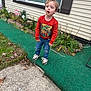 child, boy, orange_sweatshirt, dinosaur_print, jeans, sneakers, sidewalk, green_carpet, garden, plants, flowers, house, window, shutters, outdoor, grass, concrete, expression, curiosity, casual_clothing