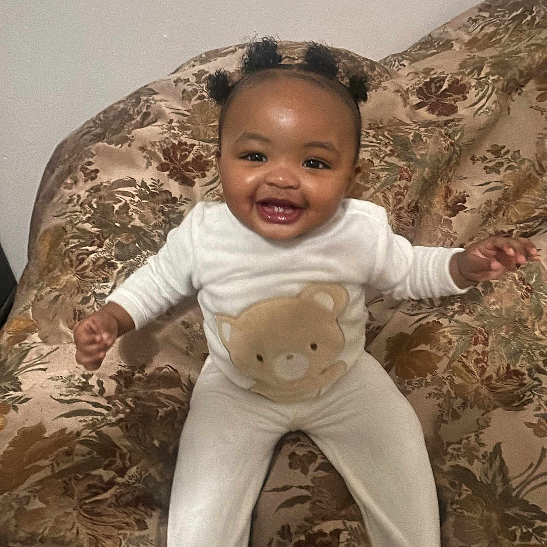Khyla joined the competition — help win amazing prizes! baby, child, couch, cozy, cute, expression, face, floral_pattern, furniture, hair_puffs, happy, indoors, infant, person, portrait, sitting, smiling, teddy_bear, white_outfit, young_child