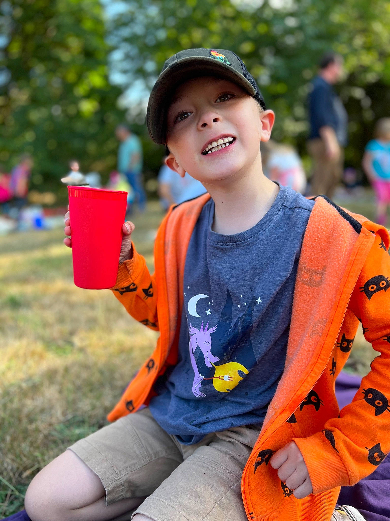 Tripp is registered to the contest to win money with this photo: bag, baseball_cap, cap, child, electric_blue, event, fun, grass, green, happy, headwear, leisure, orange, person, personal_protective_equipment, recreation, sitting, smile, sportswear, t_shirt