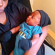 Carter is registered to the contest to win money with this photo: baby, adult, holding, blue_onesie, black_hat, indoor, person, face, arm, clothing, infant, curious, close_up, skin, seated, interaction, parent, child, casual, expression