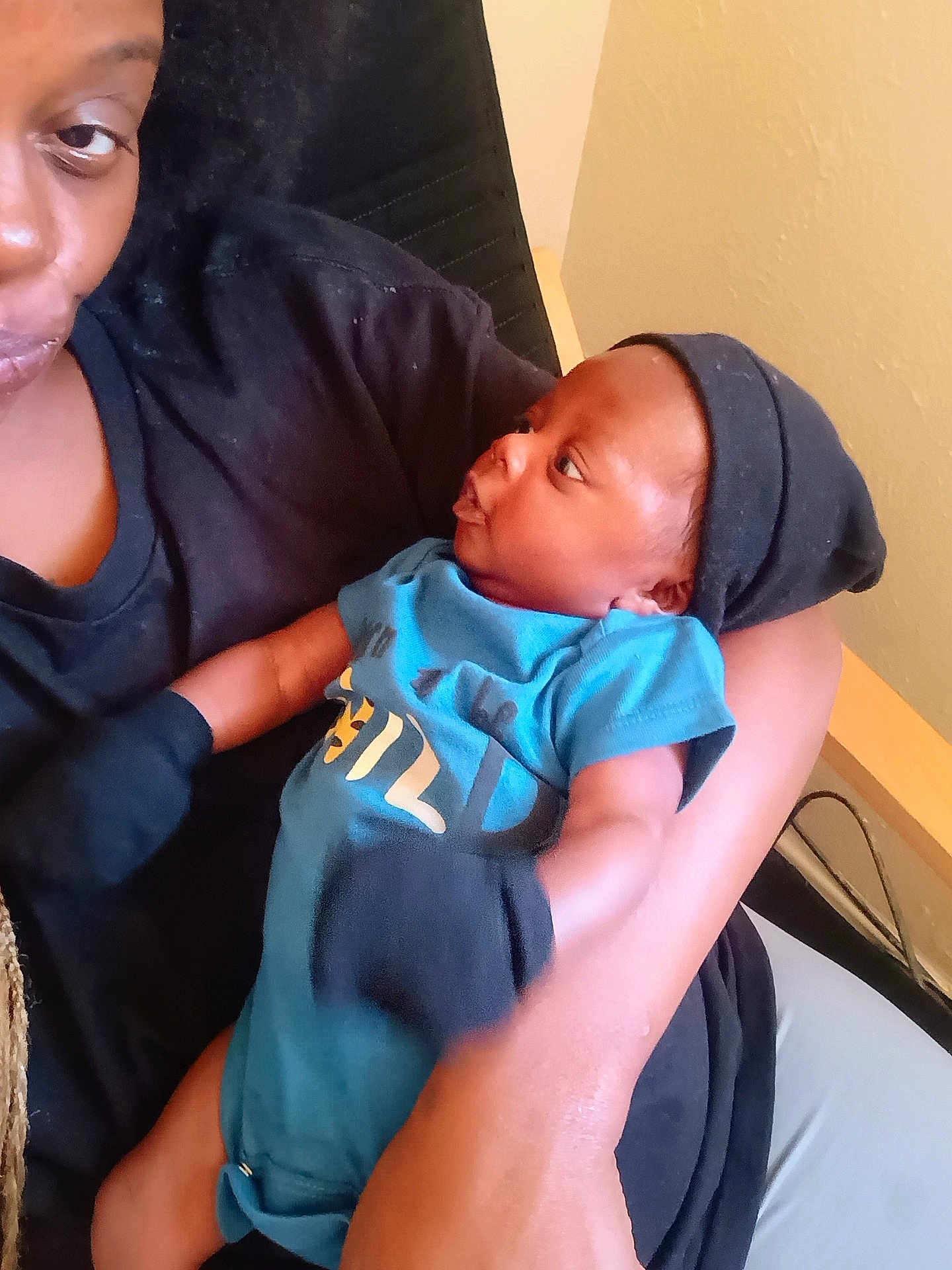 Carter is registered to the contest to win money with this photo: baby, adult, holding, blue_onesie, black_hat, indoor, person, face, arm, clothing, infant, curious, close_up, skin, seated, interaction, parent, child, casual, expression