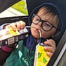 child, glasses, hoodie, juice_box, car_seat, seatbelt, window, drinking, multitasking, casual_clothing, daylight, indoors, person, face, young_child, cute, playful, seat, relaxing, funny