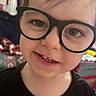 child, toddler, glasses, smile, face, closeup, indoors, black_shirt, teeth, happy, cute, portrait, young, eyewear, person, head, skin, hair, expression, baby
