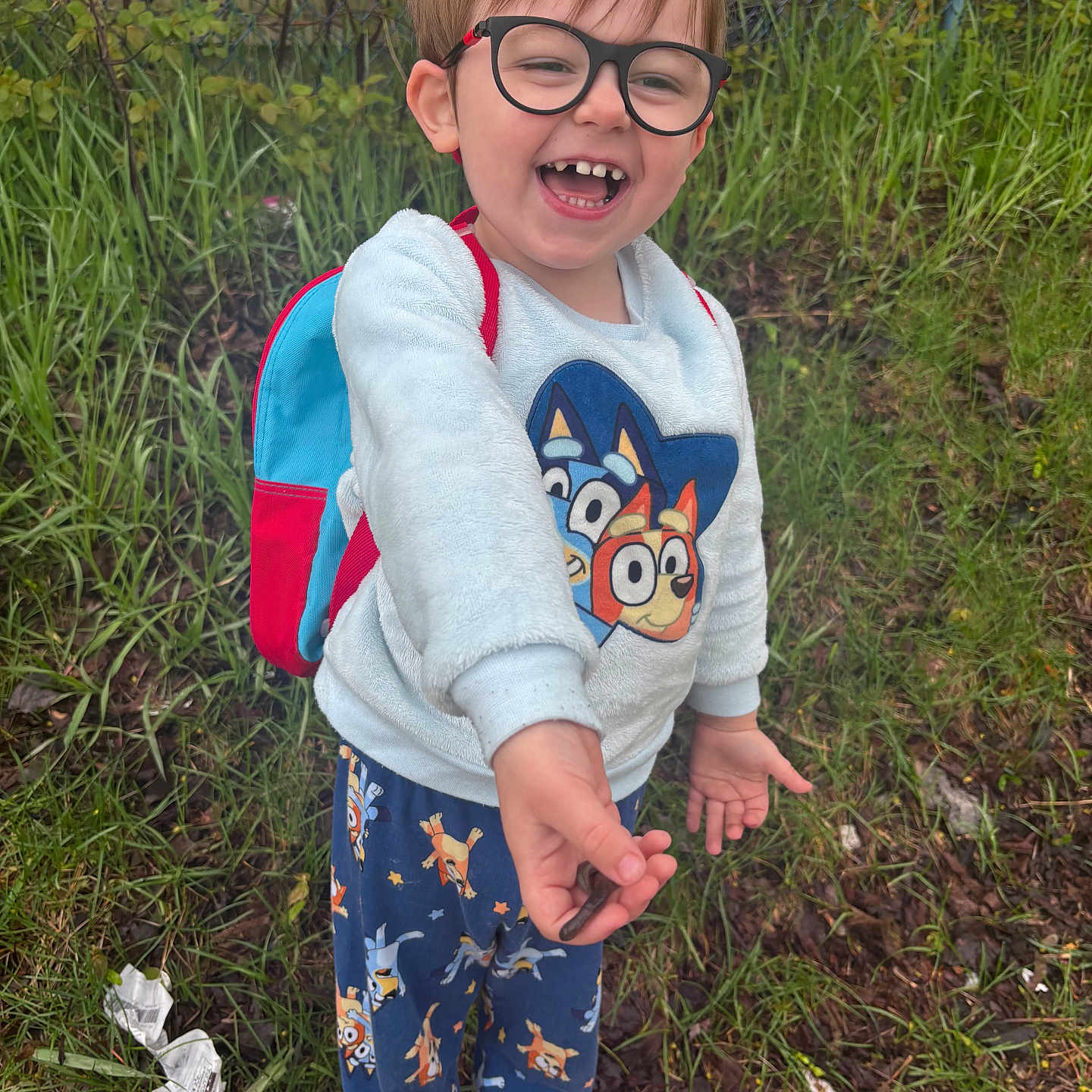 Greyson is registered to the contest to win money with this photo: child, smiling, glasses, backpack, outdoor, grass, fence, greenery, sweater, pajama_pants, footwear, happy, nature, person, young, standing, playful, cute, casual_clothing, holding_object