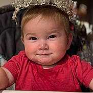Emilia joined the competition — help win amazing prizes! baby, child, crown, high_chair, red_shirt, smile, face, portrait, indoor, cute, infant, jewelry, headwear, person, closeup, young_child, happy, seated, adorable, fashion_accessory
