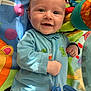 baby, smiling, onesie, blue_clothing, play_mat, toy, colorful, infant, happy, face, child, crawling_mat, cute, indoor, person, young_child, dinosaur_print, bright_colors, soft_toys, closeup
