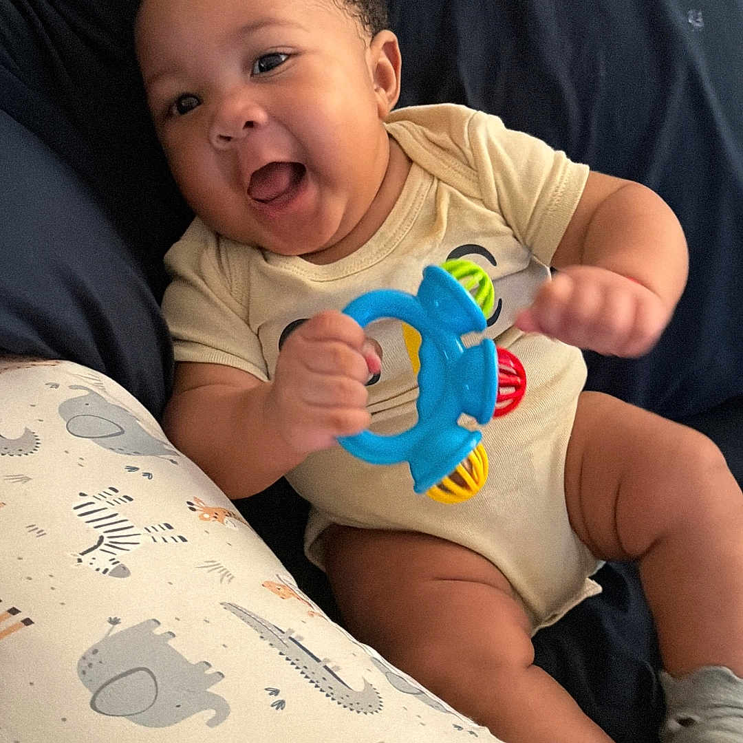 Jon-Michael is registered to the contest to win money with this photo: baby, infant, child, smiling, laughing, sitting, portrait, indoor, cushion, blanket, onesie, rattle, toy, colorful_toy, bed, nursery, animal_print, happy, face, photography