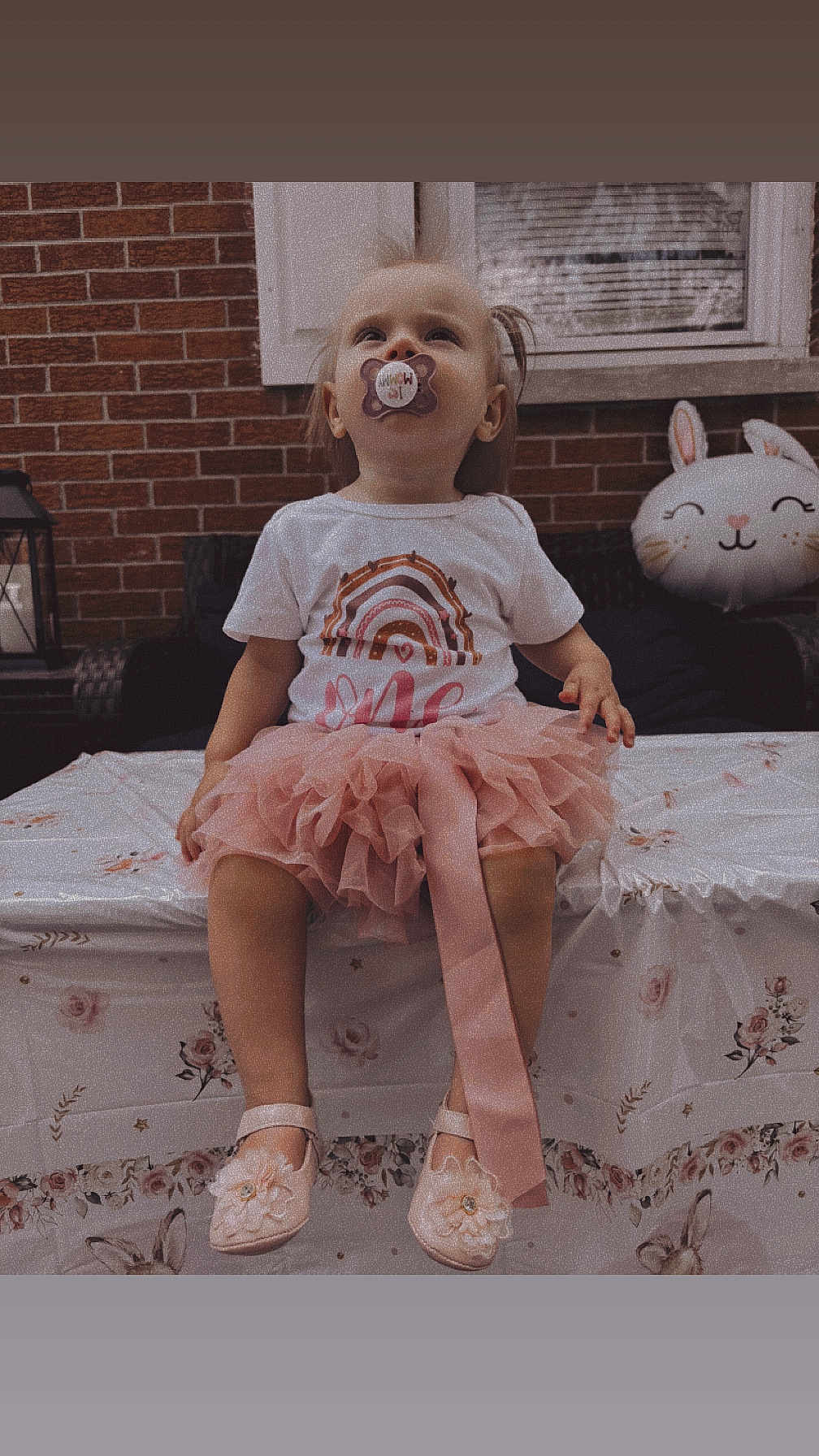 Oaklynn is registered to the contest to win money with this photo: toddler, child, pacifier, pink_tutu, shoes, table, tablecloth, brick_wall, window, balloon, bunny, outdoor, curious, sitting, face, cute, clothing, footwear, decor, party