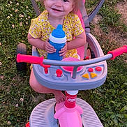 Oaklynn is registered to the contest to win money with this photo: toddler, child, smiling, tricycle, toy, outdoor, grass, yellow_clothing, pink, blue, sippy_cup, happy, baby, playtime, summer, daylight, feet, seat, handlebars, nature