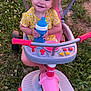 toddler, child, smiling, tricycle, toy, outdoor, grass, yellow_clothing, pink, blue, sippy_cup, happy, baby, playtime, summer, daylight, feet, seat, handlebars, nature