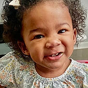 Milan is registered to the contest to win money with this photo: toddler, child, smiling, curly_hair, bow, floral_clothing, indoor, happy, portrait, person, cute, baby_teeth, face, young_child, home, furniture, plant, shirt, standing, light