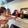 baby, child, elf_on_the_shelf, toy, fireplace, mantel, figurine, plush_toy, person, hand, wall, photo_frame, decor, indoor, curious, reaching, toddler, candle, ceramic, furniture