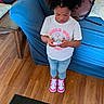 child, girl, standing, indoor, playing, gadget, white_tshirt, jeans, pink_sneakers, wooden_floor, blue_couch, hair, focused, casual_clothing, young, person, floor, furniture, technology, home