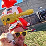 man, dog, selfie, outdoor, inflatable, red_fox, guitar, sunglasses, hat, grass, building, blue_sky, daylight, smile, pet, casual_clothing, festival, event, fun, sunny