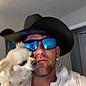 man, dog, indoor, cowboy_hat, sunglasses, white_dog, pet, shoulder, casual, portrait, facial_hair, reflection, clothing, closeup, human, companion, animal, headwear, indoor_lighting, selfie