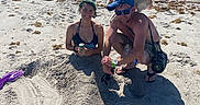 Goldie joined the competition — help win amazing prizes! beach, sand, woman, man, dog, sunny, blue_sky, seaweed, smiling, sunglasses, hat, casual_clothing, outdoor, vacation, relaxation, playful, buried_in_sand, pet, shadow, fun