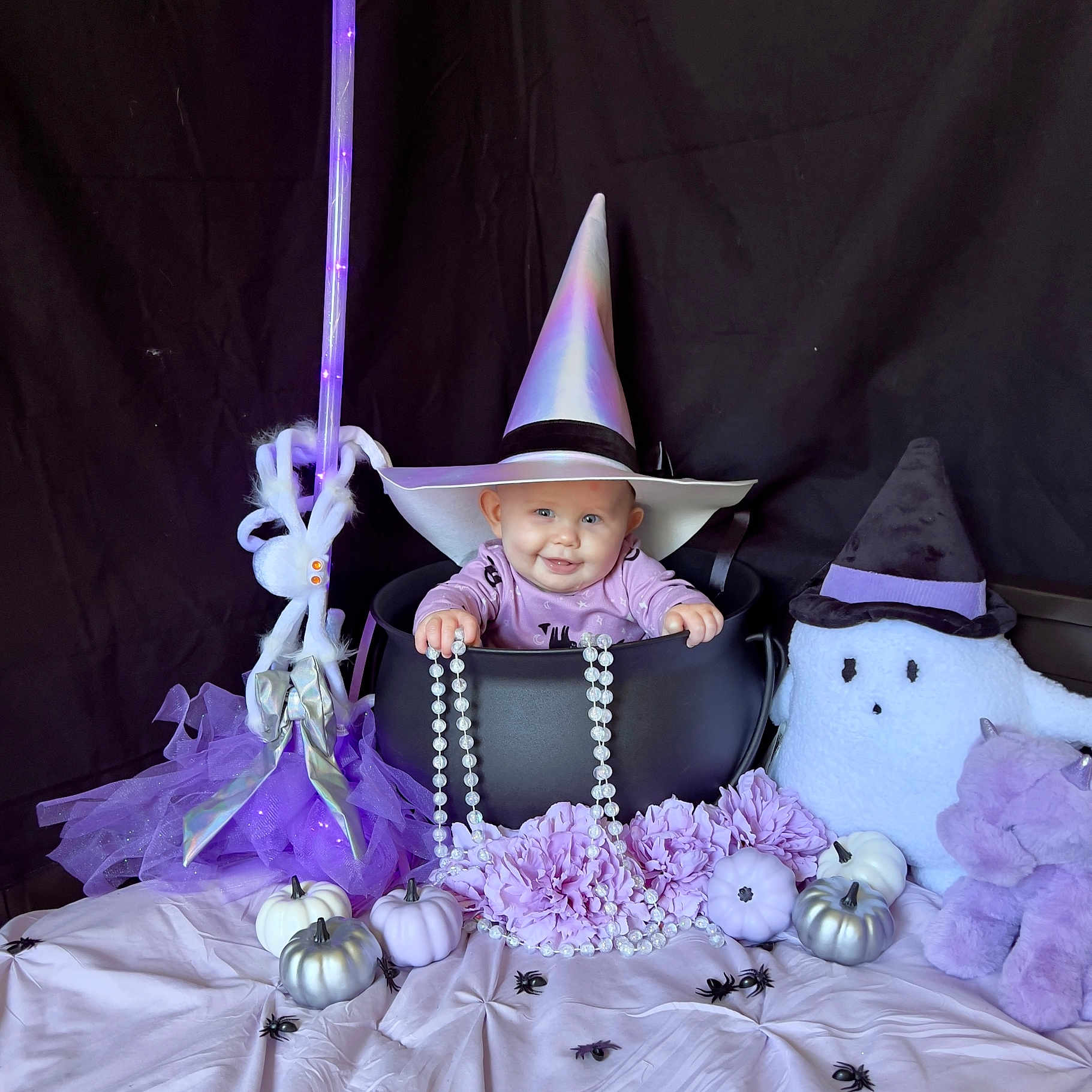 Kameron is registered to the contest to win money with this photo: baby, baby_smiling, beads, black_background, broomstick, cauldron, costume, cute, decor, flowers, ghost_toy, halloween, indoor, plush_toy, portrait, pumpkin, purple, spiders, unicorn_toy, witch_hat