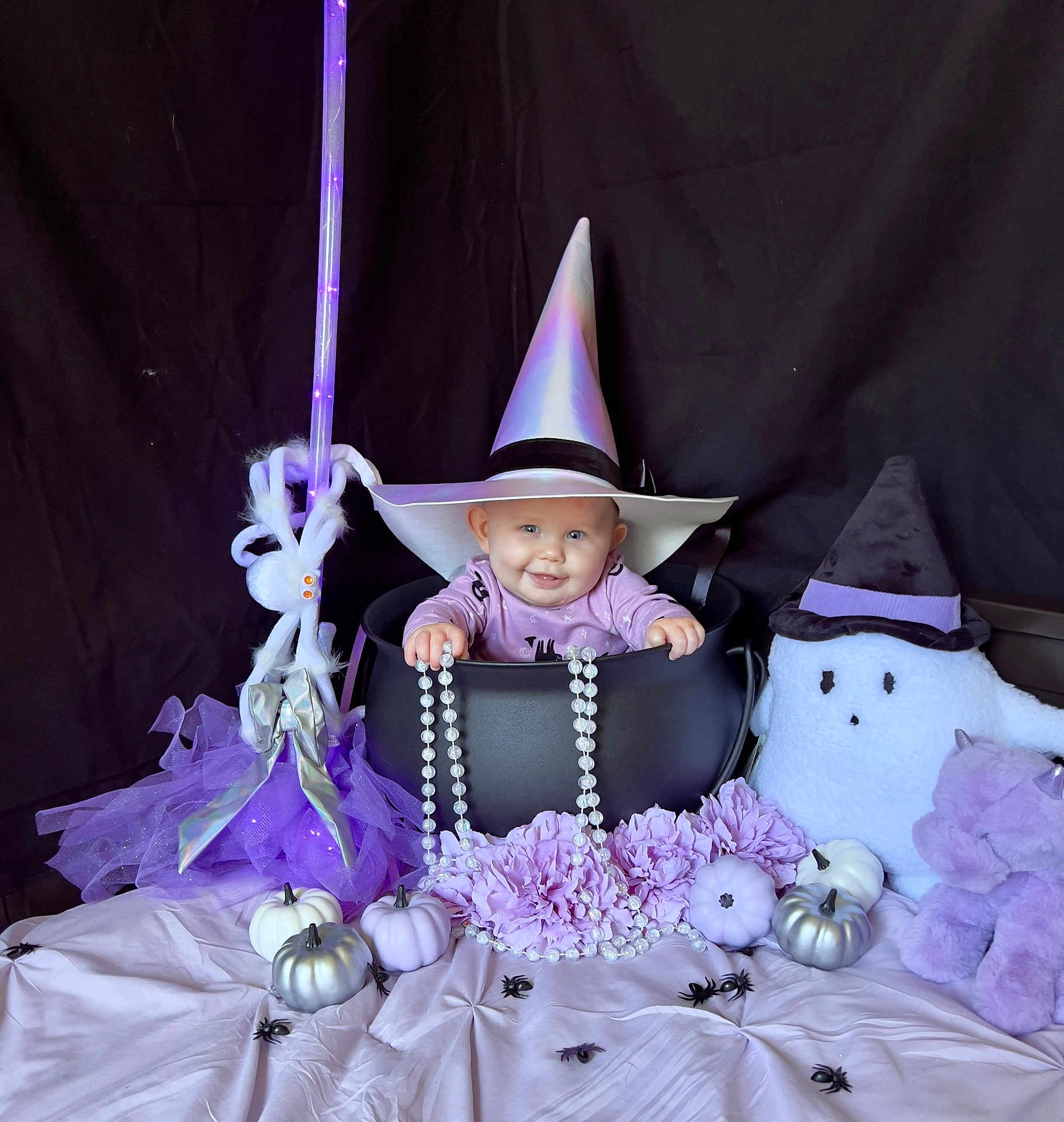 Kameron is registered to the contest to win money with this photo: baby, witch_hat, cauldron, purple, black_background, plush_toy, ghost_toy, unicorn_toy, pumpkin, decor, spiders, broomstick, flowers, beads, costume, baby_smiling, halloween, cute, indoor, portrait