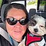 woman, dog, pet, car_interior, selfie, sunglasses, seatbelt, red_harness, small_dog, fluffy_dog, hand, face, portrait, casual, daylight, vehicle, seat, window, relaxed, smile
