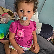 Sapphire is registered to the contest to win money with this photo: toddler, child, pacifier, curly_hair, pink_shirt, flowers, sitting, couch, stuffed_toys, plush, indoor, young_child, portrait, cute, looking_at_camera, colorful, play_area, comfort, casual_clothing, baby