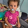 toddler, child, pacifier, curly_hair, pink_shirt, flowers, sitting, couch, stuffed_toys, plush, indoor, young_child, portrait, cute, looking_at_camera, colorful, play_area, comfort, casual_clothing, baby