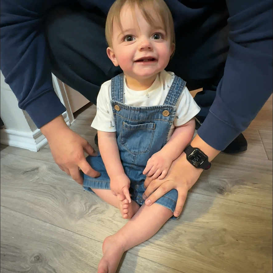 Noah is registered to the contest to win money with this photo: toddler, child, denim_overalls, smile, floor, wooden_floor, adult, hands, watch, casual_clothing, indoor, person, baby, sitting, happy, light_skin, short_hair, white_tshirt, support, bonding