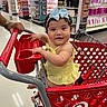 baby, child, shopping_cart, target, store, retail, grocery, person, toddler, yellow_dress, blue_bow, hand, floor, shelves, products, cosmetics, smile, happy, indoor, shopping