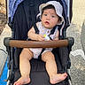 baby, child, stroller, outdoor, hat, striped_clothing, toy, pavement, sunhat, footwear, seatbelt, curious_expression, infant, summer, park, shade, baby_clothes, cute, sitting, person