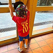 Thiago a rejoint le concours — aidez-le/la à gagner de superbes lots ! child, kid, back_view, red_jersey, espana, number_10, cap, superhero_design, glass_door, door_handle, indoor, tiled_floor, blue_shorts, sandals, outdoor_view, car, greenery, window, standing, reaching