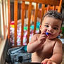 baby, bedding, chewing, child, colorful, crib, curious, curly_hair, expression, face, hand, indoor, person, room, skin, toddler, toy, window_light, wooden_furniture, young