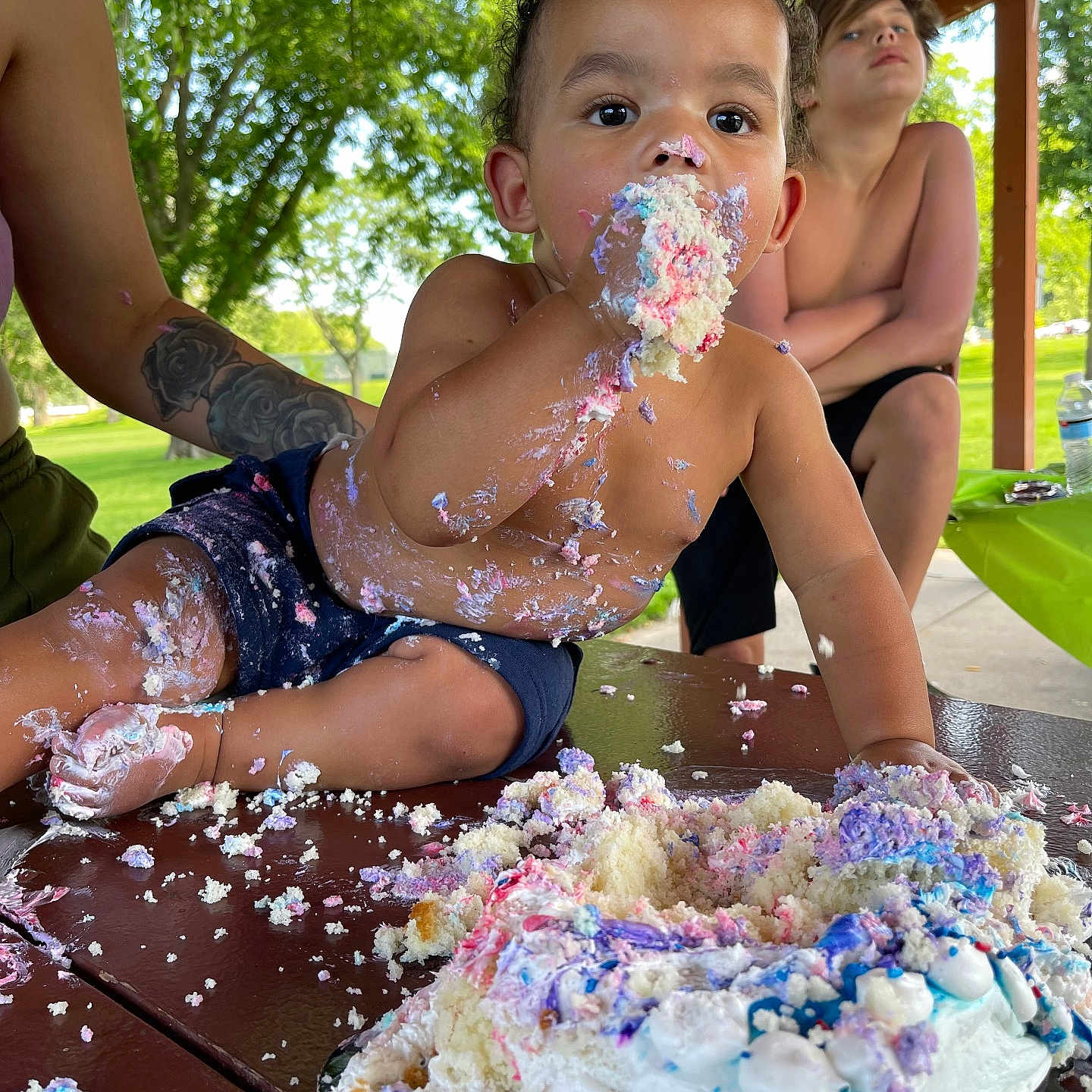 Vayden is registered to the contest to win money with this photo: birthday_cake, cake, celebration, child, daylight, family, food, frosting, fun, messy, naked_torso, outdoor, park, people, person, picnic_table, smiling, summer, tattoo, toddler