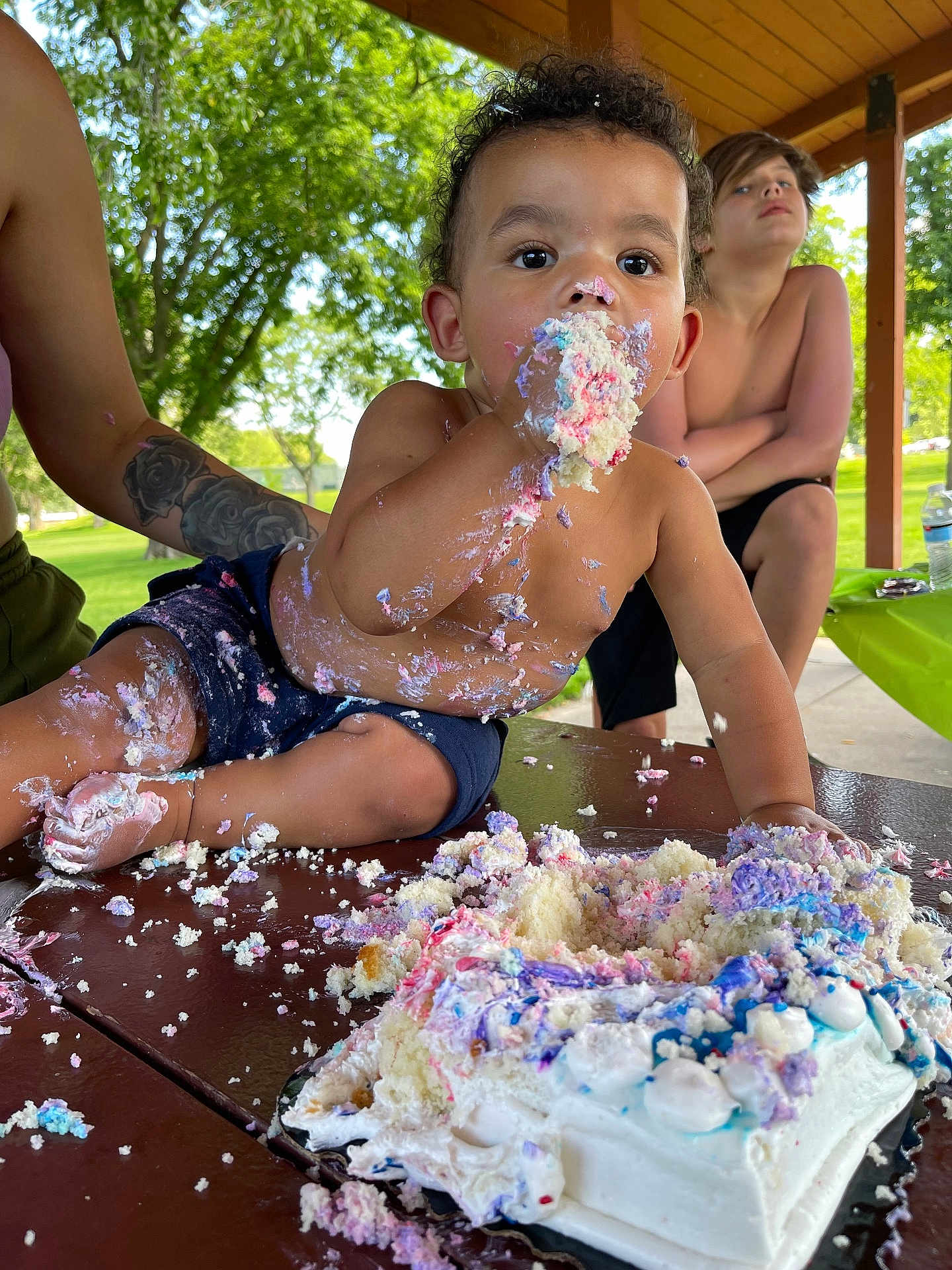 Vayden is registered to the contest to win money with this photo: birthday_cake, cake, celebration, child, daylight, family, food, frosting, fun, messy, naked_torso, outdoor, park, people, person, picnic_table, smiling, summer, tattoo, toddler